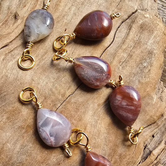 Four teardrop-shaped gemstone pendants with gold wire loops, arranged on a textured piece of natural wood. The stones feature marbled patterns in shades of brown, purple, and gray.